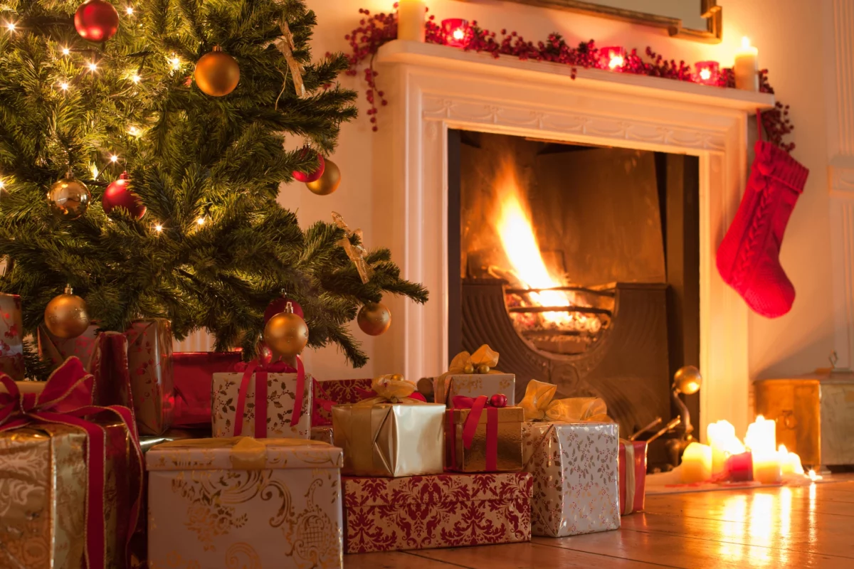 Christmas Tree and Presents Next to a lit fire place with a stocking handing down from the fire place mantle.